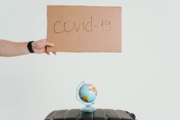 A hand holding a cardboard sign with the writing "covid-19" written on it, above a small coloured globe on a surface