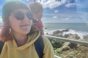 Millie, a woman with half black and half red hair, wearing a green beanie, sunglasses and a green hat is baby wearing TinyHuman, a one year old boy with long hair tied up into a bun, as they stand next to a railing overlooking the ocean.